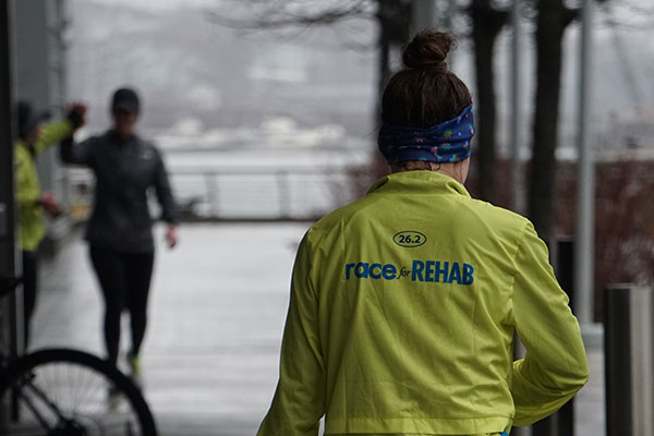 A runner in a yellow Race for Rehab jacket, seen from behind.