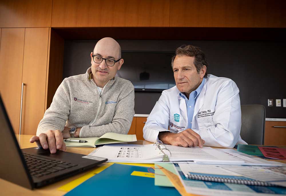 Two researchers sit at a desk cluttered with papers and binders, reviewing something on a laptop.