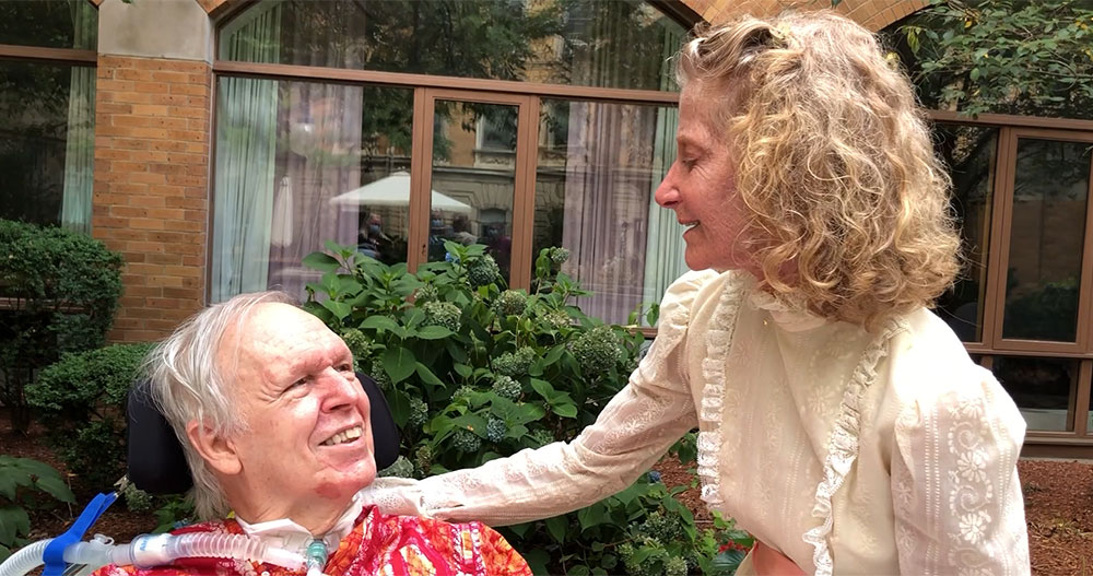Randy, an older man in a wheelchair, and Catherine, an older woman in a white dress, share a moment on their patio.