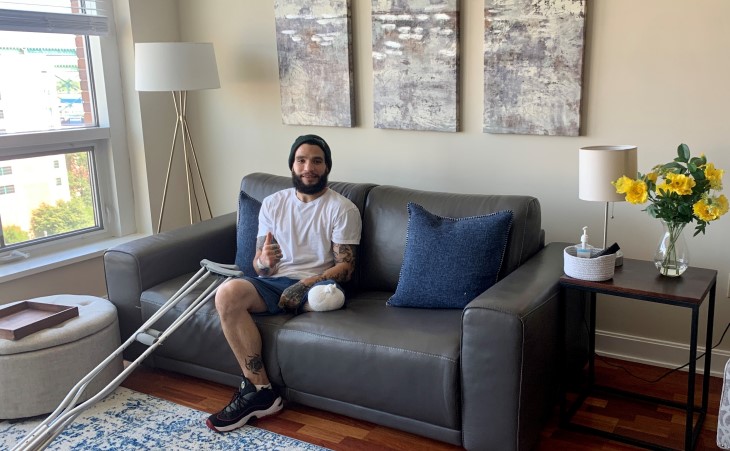 Chris giving a thumbs-up on his sofa with his crutches beside him.