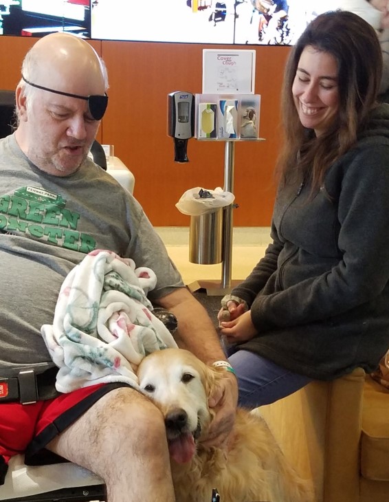 John sitting in a chair with a golden retriever leaning against his leg, and his daughter sitting on the arm of another chair and smiling at the dog.