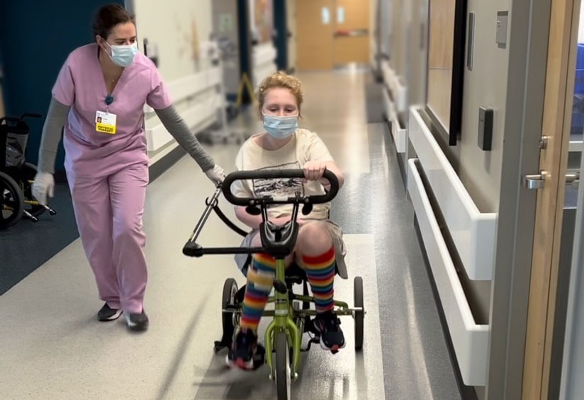 A 12-year-old girl in rainbow socks cycles an adaptive tricycle down a hospital hallway, assisted by her physical therapist.