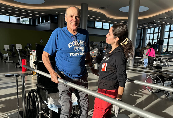 Sandy, an older man, doing a walking exercise at the parallel bars with his physical therapist.