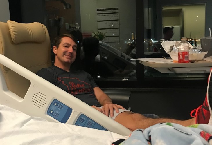 Tim looking cheerful in a chair beside his hospital bed.